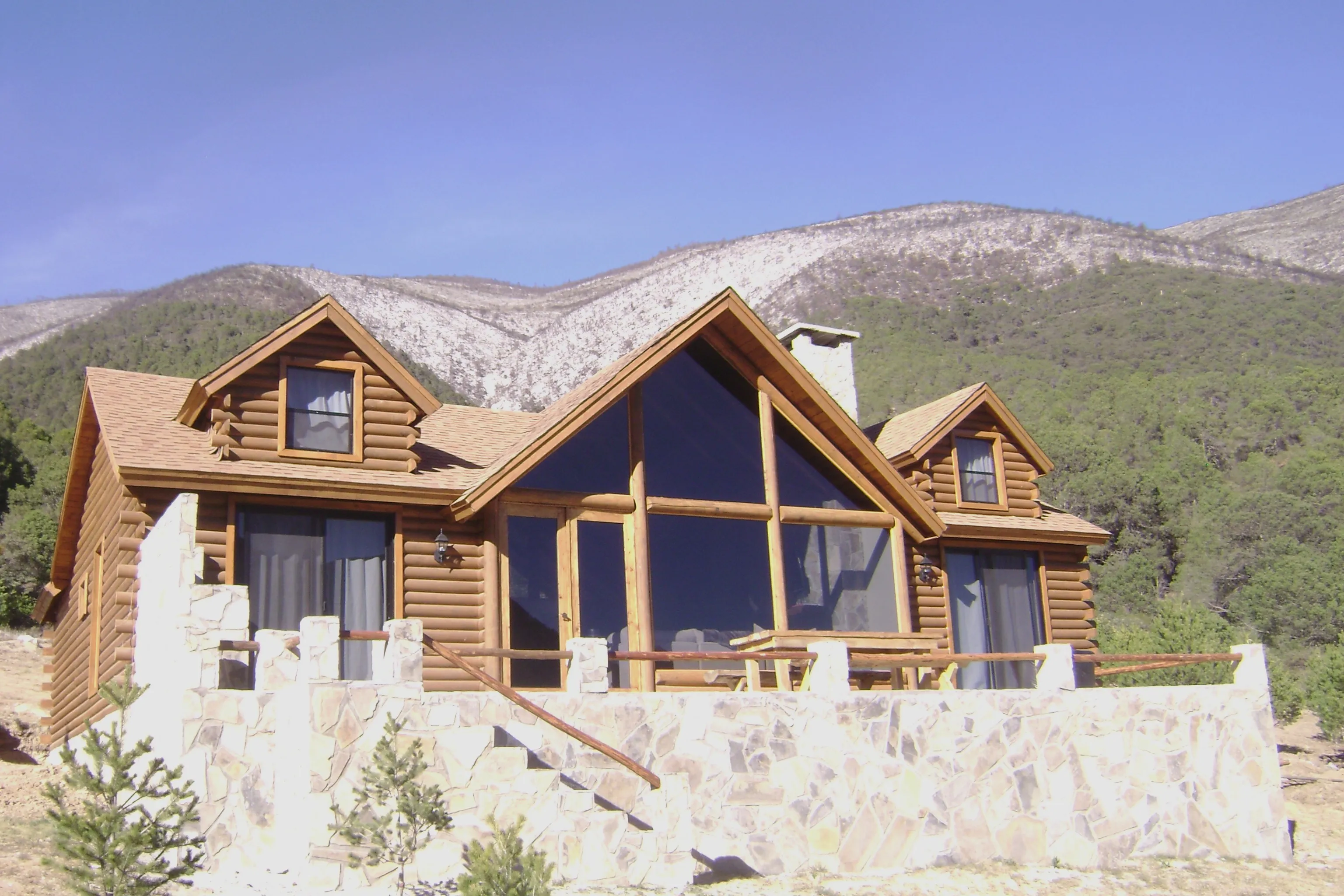 Cabaña de tronco con fachada de ventanal frontal, construida sobre terraza de piedra al pie de la sierra